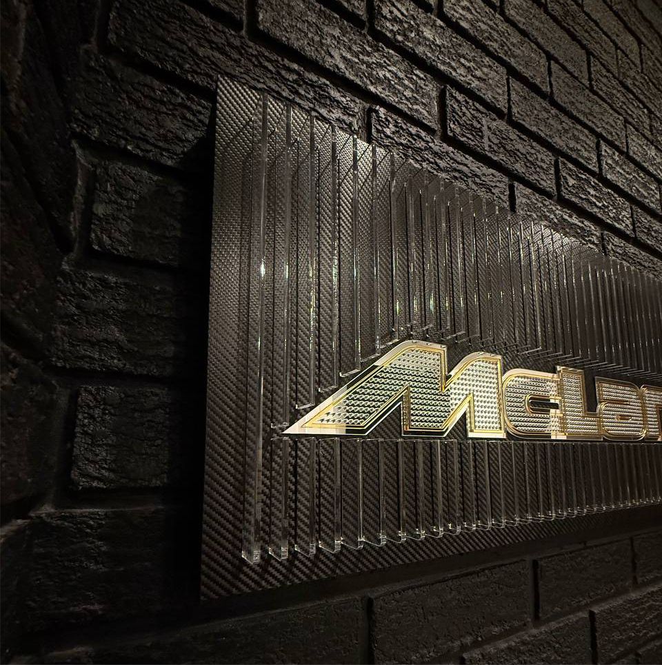 Full display shot of the McLaren-inspired acrylic artwork with layered fins, micro-logo texture, carbon-fiber background, and illuminated orange LED lighting.