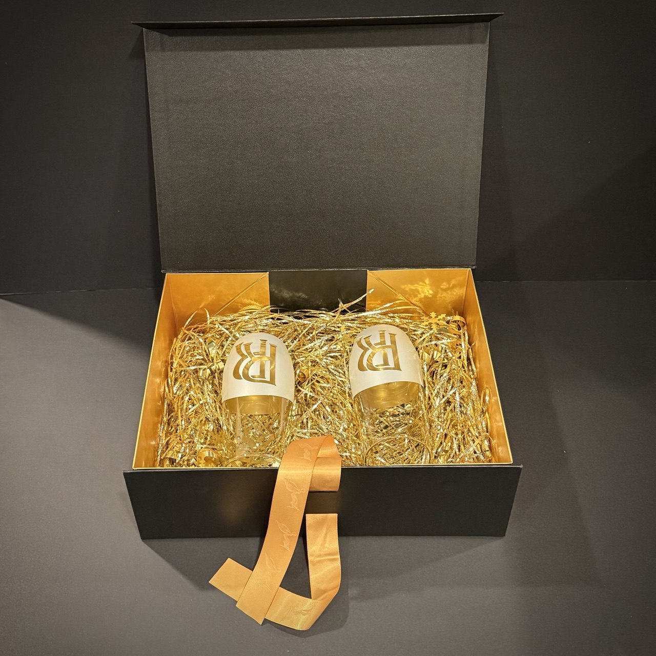 Open premium black gift box displaying silver and gold spirit-inspired glasses arranged with gold decorative filler