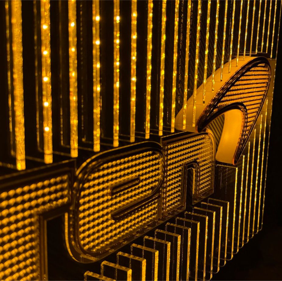 Angled view of a McLaren-inspired illuminated wall piece with stacked acrylic fins, micro-logo patterns, carbon-fiber background, and warm orange lighting.