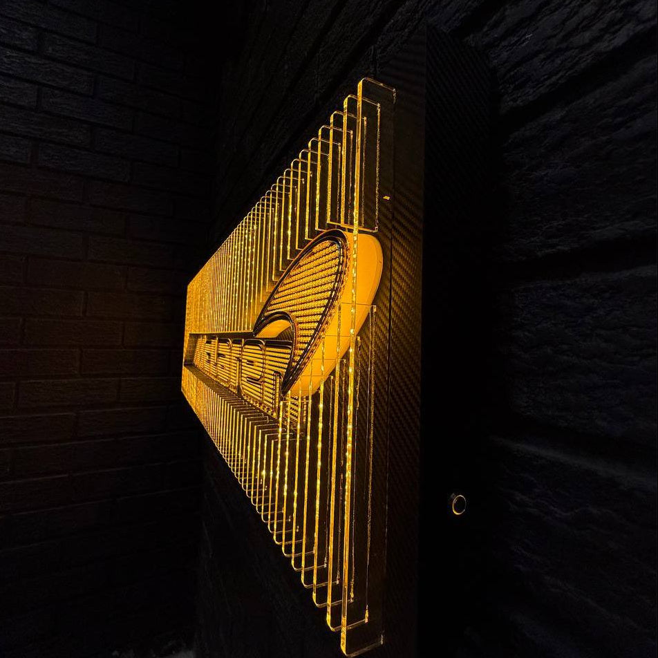 Artistic shot of a McLaren-inspired illuminated acrylic display with stacked fins, carbon-fiber backing, and micro-logo texture illuminated in warm orange tones.