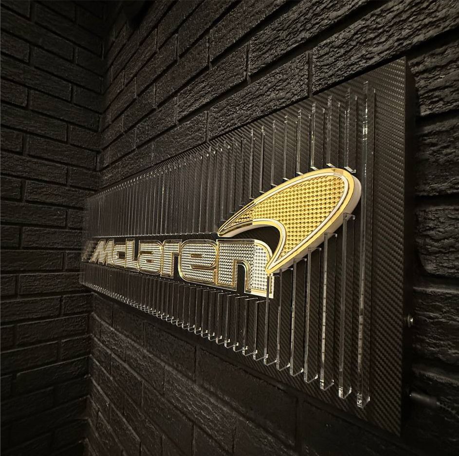 Side-angle photo of a McLaren-inspired illuminated acrylic structure featuring micro-logo details, carbon-fiber backing, and bright orange LED highlights.