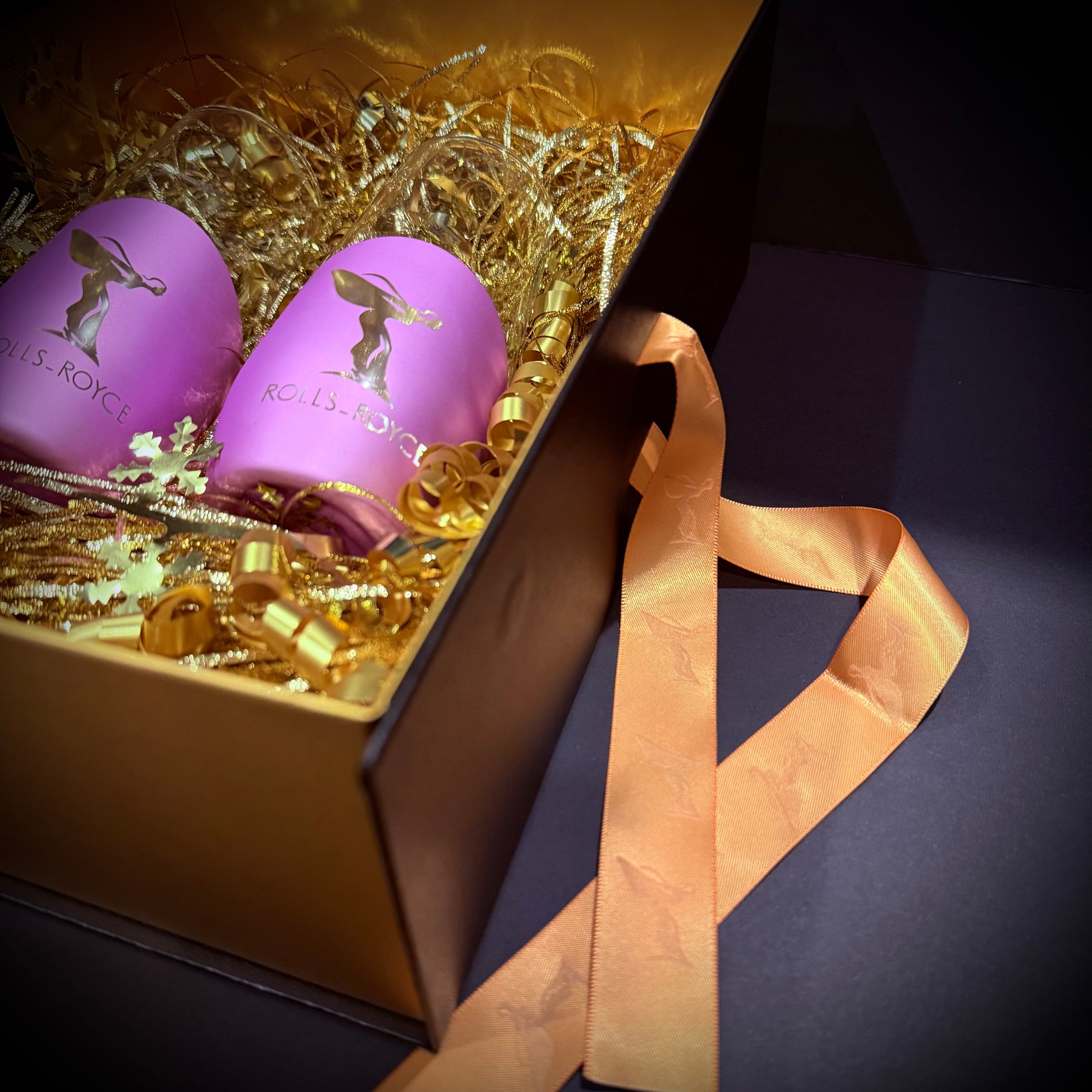 Gift box containing two purple luxury glasses with gold decorative ribbon and shredded filler.