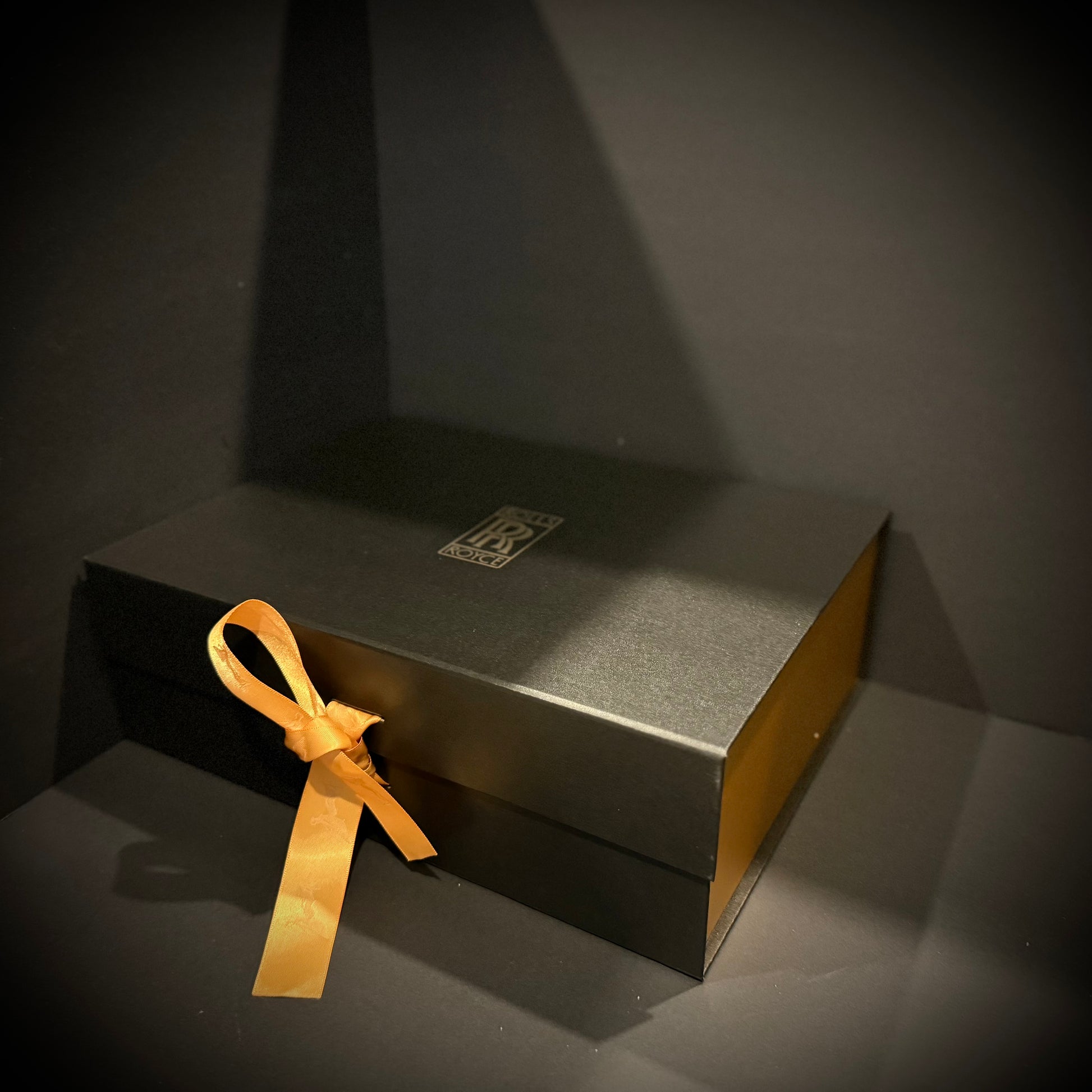 Black luxury gift box with a gold ribbon and subtle RR-inspired emblem on top.